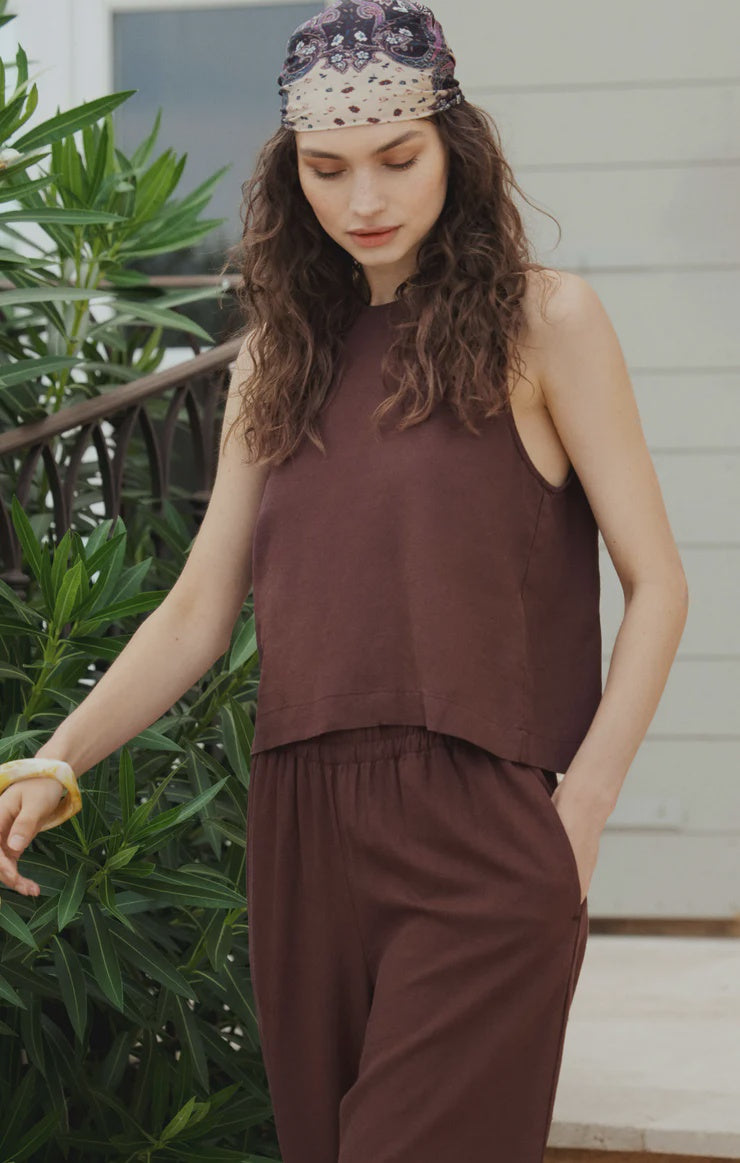 A woman with wavy brown hair stands outdoors near green plants and a railing, wearing the Zertz SLOANE LINEN TOP—a relaxed fit sleeveless brown linen blend tank—with matching pants, one hand in her pocket.