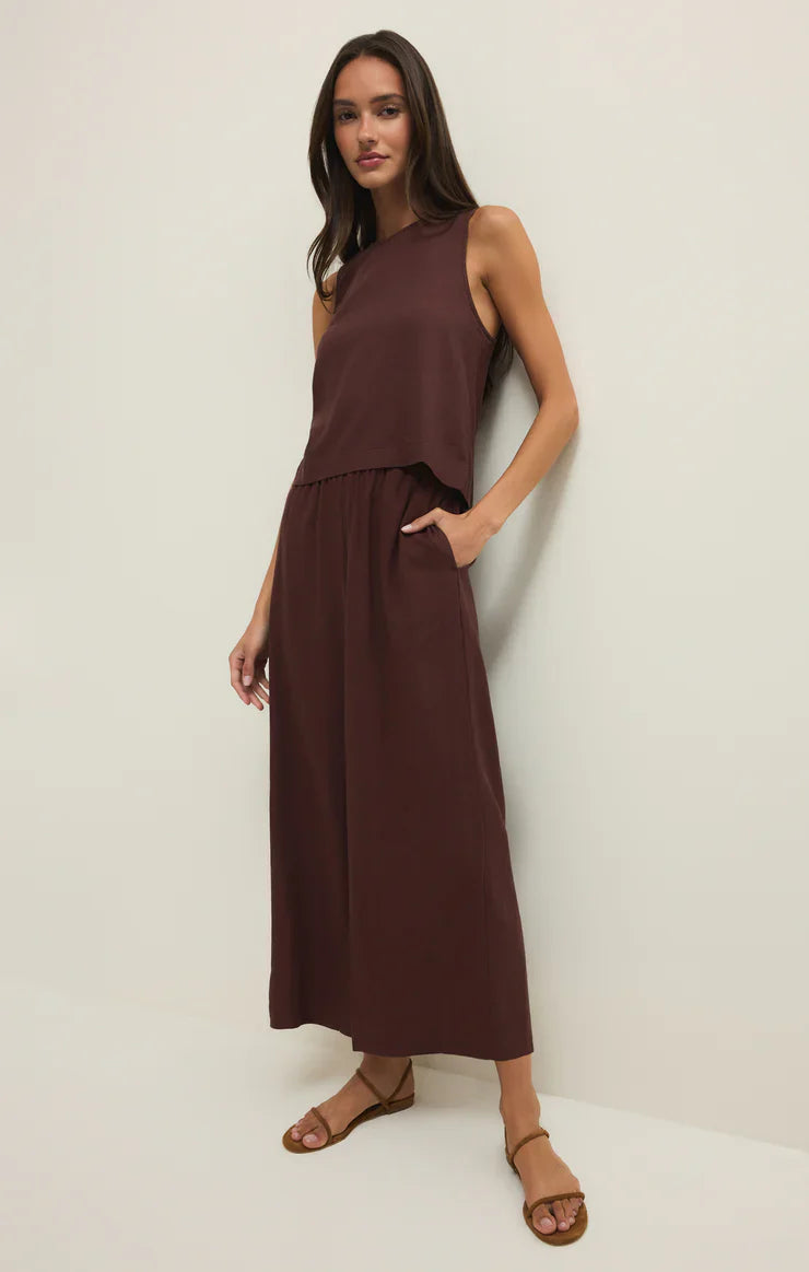 A woman wearing the Zertz SLOANE LINEN TOP, a sleeveless dark brown piece with a relaxed fit, paired with brown sandals, stands against a plain white wall with one hand in her pocket.