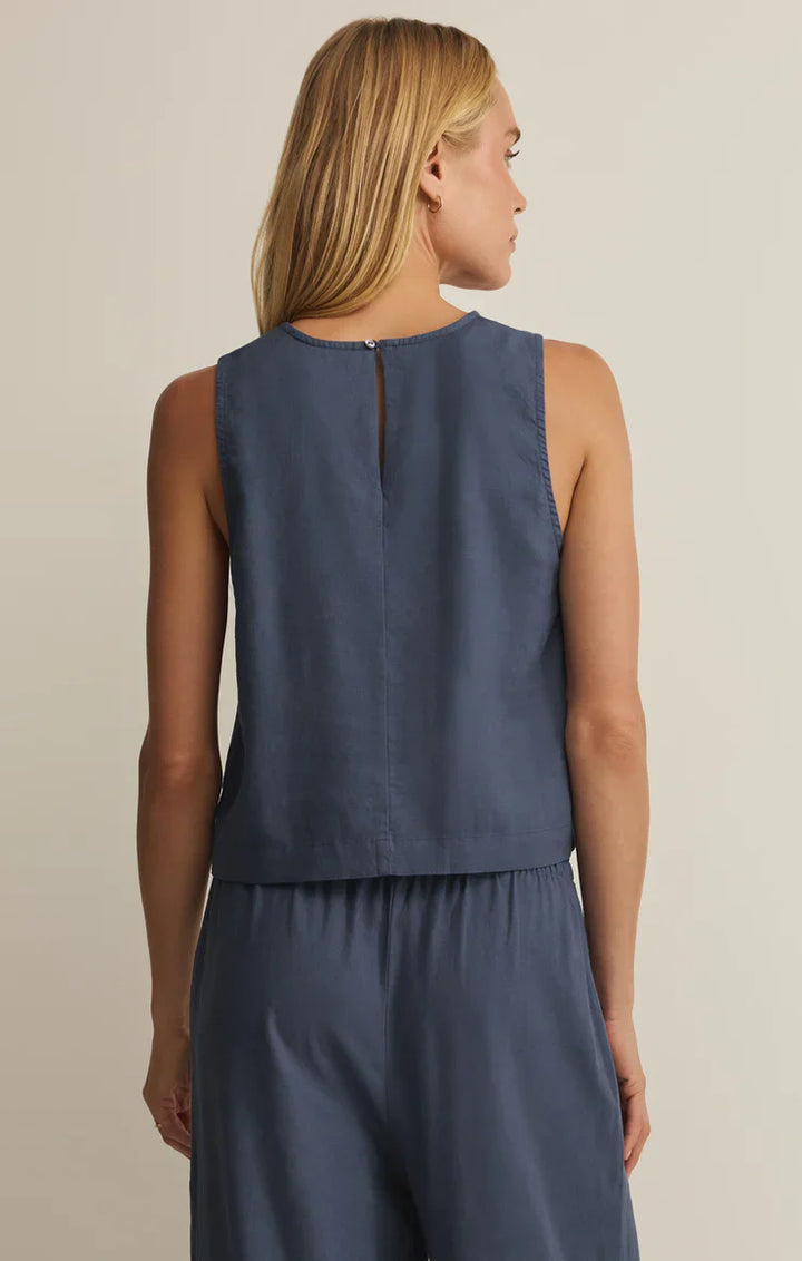 A woman with long blonde hair is shown from behind, wearing the sleeveless SLOANE LINEN TOP by Zertz in blue. The relaxed fit and button closure at the upper back flatter her silhouette as she stands against a plain light background.