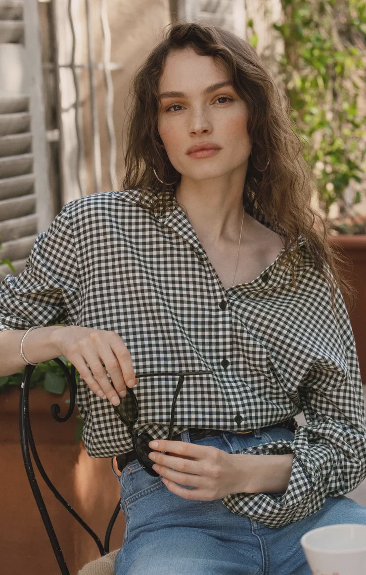 A woman with wavy brown hair sits outdoors wearing the Zertz SEAPORT BUTTON UP TOP and blue jeans. She holds sunglasses, looking calmly at the camera, with green plants and a window behind her.