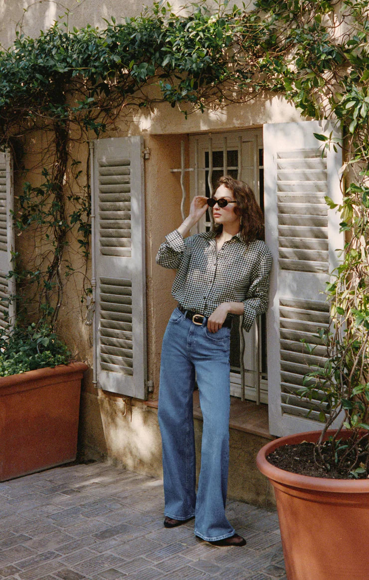 A woman in sunglasses and the Zertz SEAPORT BUTTON UP TOP with wide-leg jeans leans against a windowsill with white shutters, surrounded by large potted plants and greenery on a sunny day.