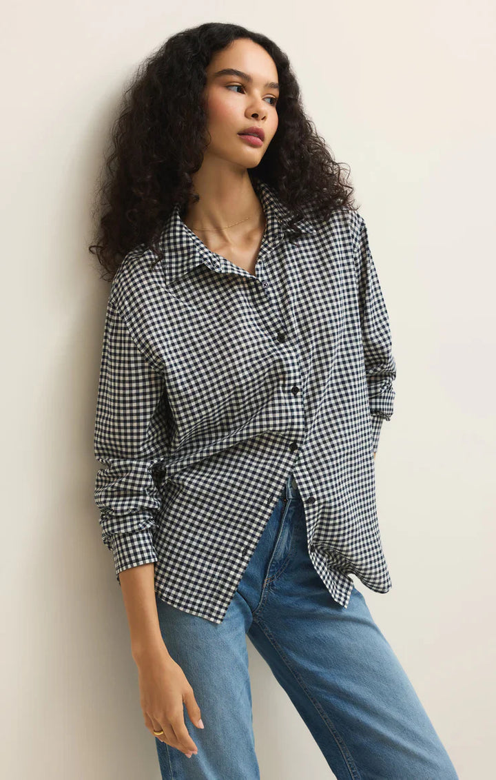A woman with long curly hair wears the Zertz SEAPORT BUTTON UP TOP in a black and white plaid print, paired with blue jeans. She leans against a light-colored wall, looking off to the side.