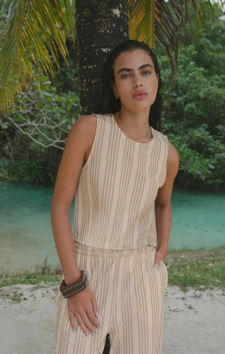 A person with long dark hair stands by turquoise water, wearing the Zertz SLOANE KNIT STRIPED TOP and a brown bracelet, against a backdrop of palm leaves and lush greenery.