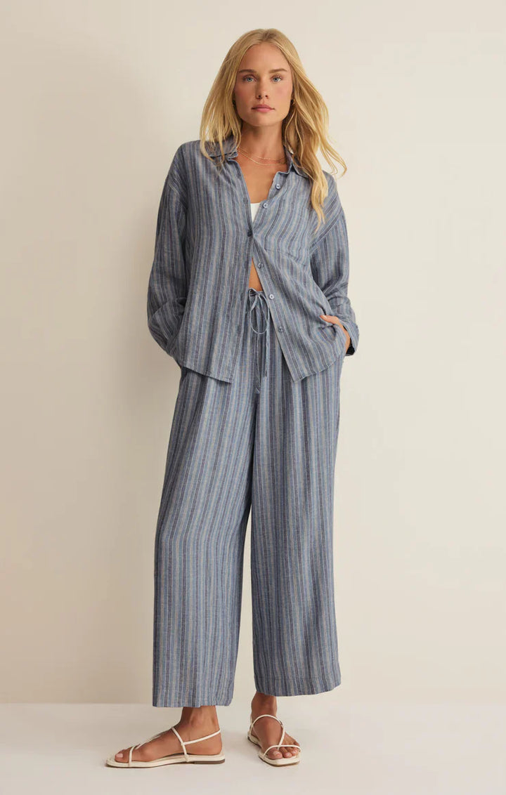 A woman stands against a plain light background, wearing Zertz’s THE PERFECT STRIPED BUTTON UP ZT262689 with matching wide-leg pants and white sandals. Her hands are in her pockets as she looks at the camera.