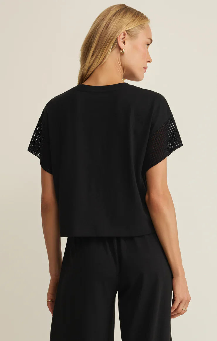 A woman with straight blonde hair wears the Zertz BANYAN TOP, a relaxed fit cotton-blend tee with mesh sleeve panels, paired with black pants and standing back to camera against a light background.