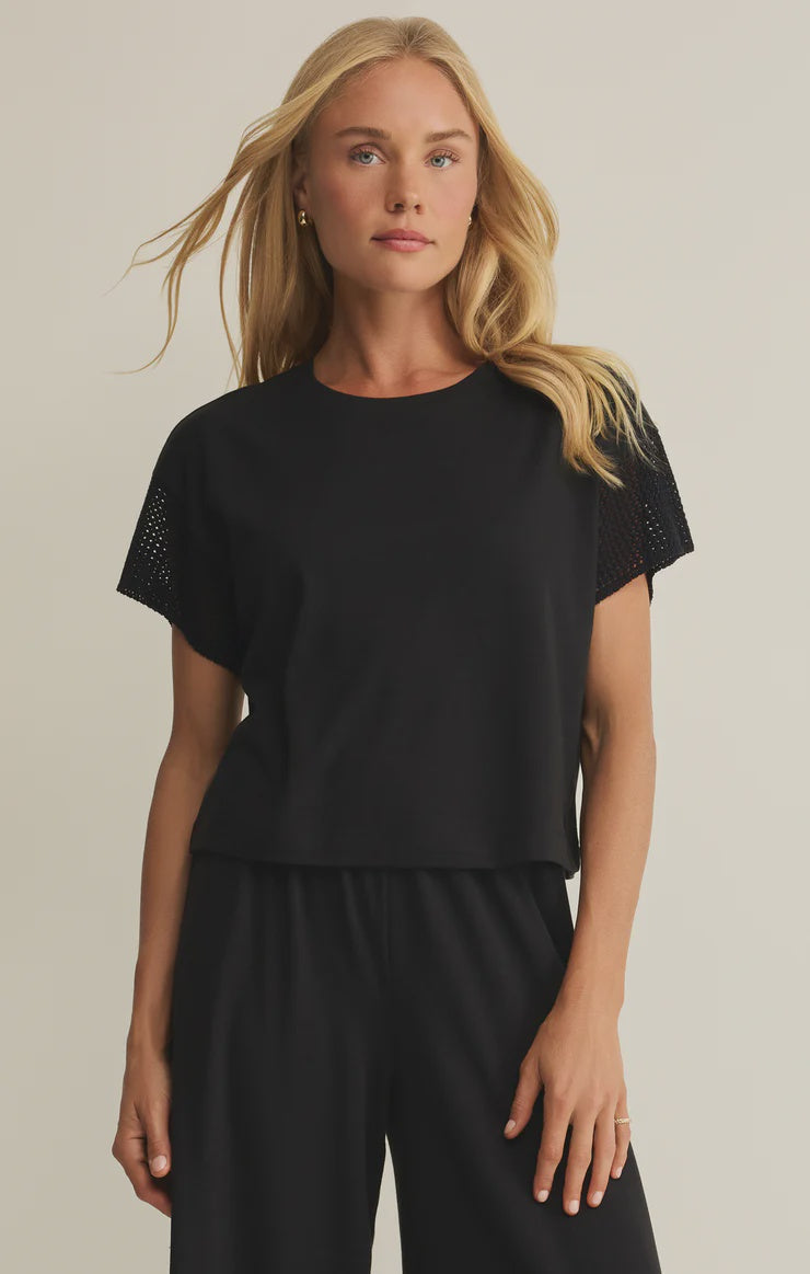 A woman with long blonde hair wears the Zertz BANYAN TOP, a relaxed fit cotton-blend t-shirt, paired with black pants and stands against a plain light background, looking straight ahead with a relaxed expression.
