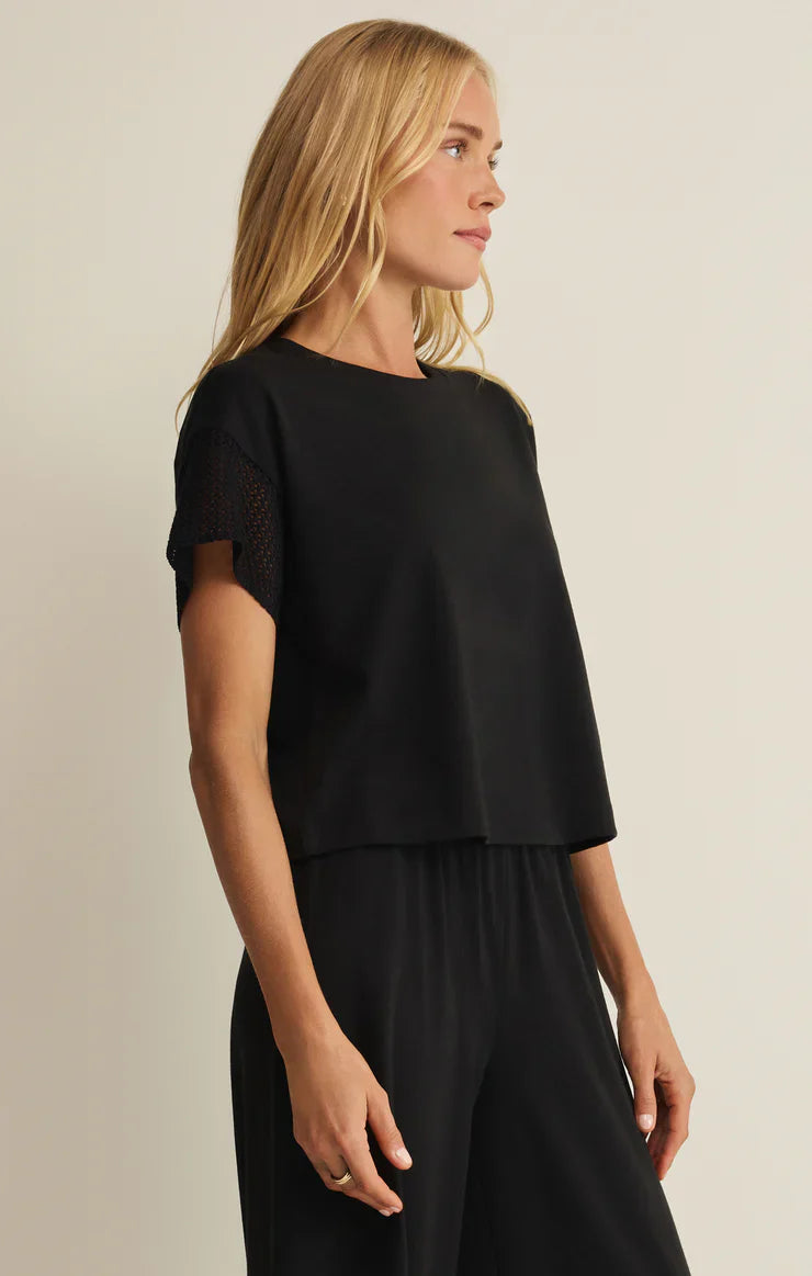 A woman with long blonde hair wears the Zertz BANYAN TOP—a relaxed-fit black cotton-blend t-shirt with crochet knit sleeves—paired with matching black pants, standing against a plain light background.
