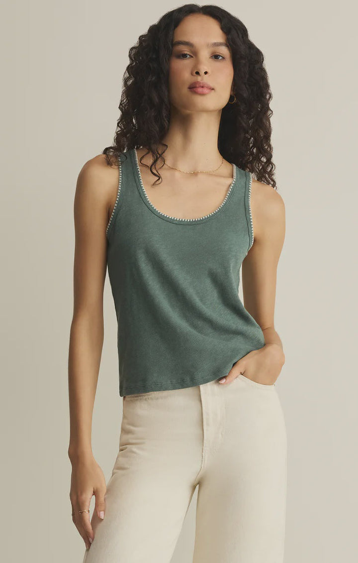 A woman with long curly hair wears the Zertz JULIAN CROCHET TANK and light-colored pants, standing against a plain beige background with one hand in her pocket and a neutral expression.