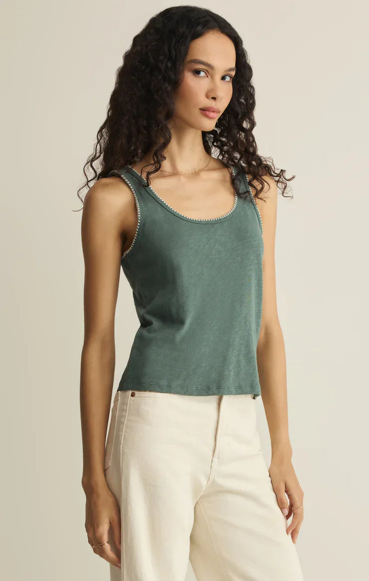 A woman with long curly hair wears the Zertz JULIAN CROCHET TANK in green and high-waisted cream pants, standing against a plain beige background.