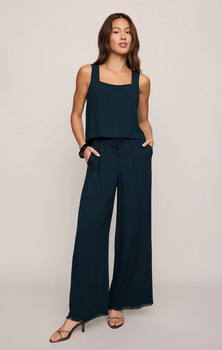 A woman stands against a neutral background wearing the Zertz CHARLI TOP, styled with wide-leg pants and heeled sandals. Her hands are in her pockets, and her loose, wavy hair frames her face.