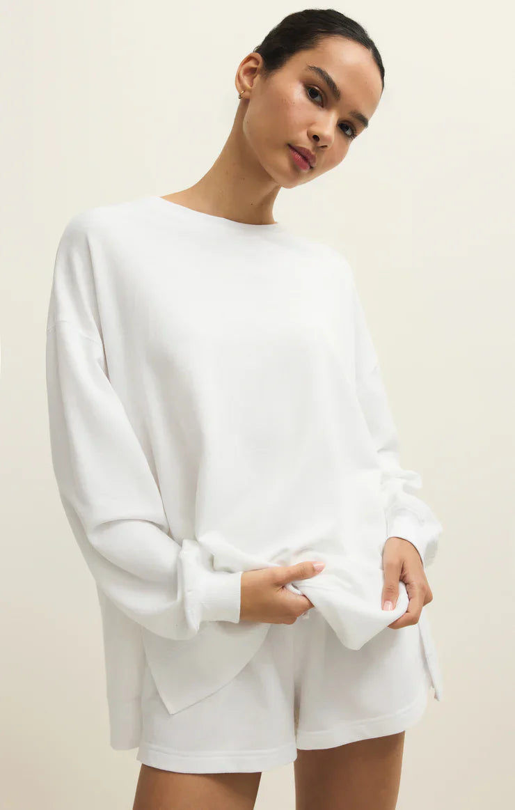 A woman stands against a plain background wearing the Zertz MODERN WEEKENDER WHITE oversized baby french terry crewneck sweatshirt with matching white shorts, looking at the camera with a neutral expression and holding the hem—a perfect weekend look.