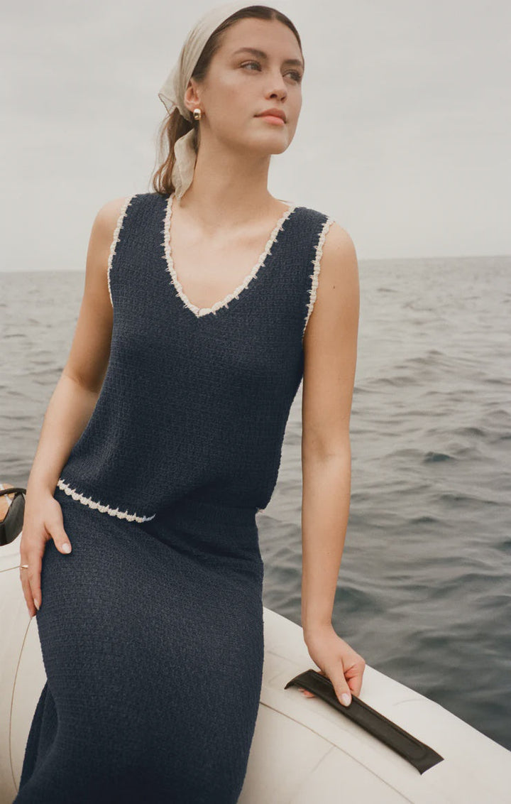 A woman in the Zertz YESSENIA WHIPSTITCH TANK, a navy blue sleeveless dress with contrast trim, sits on a boat at sea. She wears a light headscarf and gazes thoughtfully over the calm water behind her.