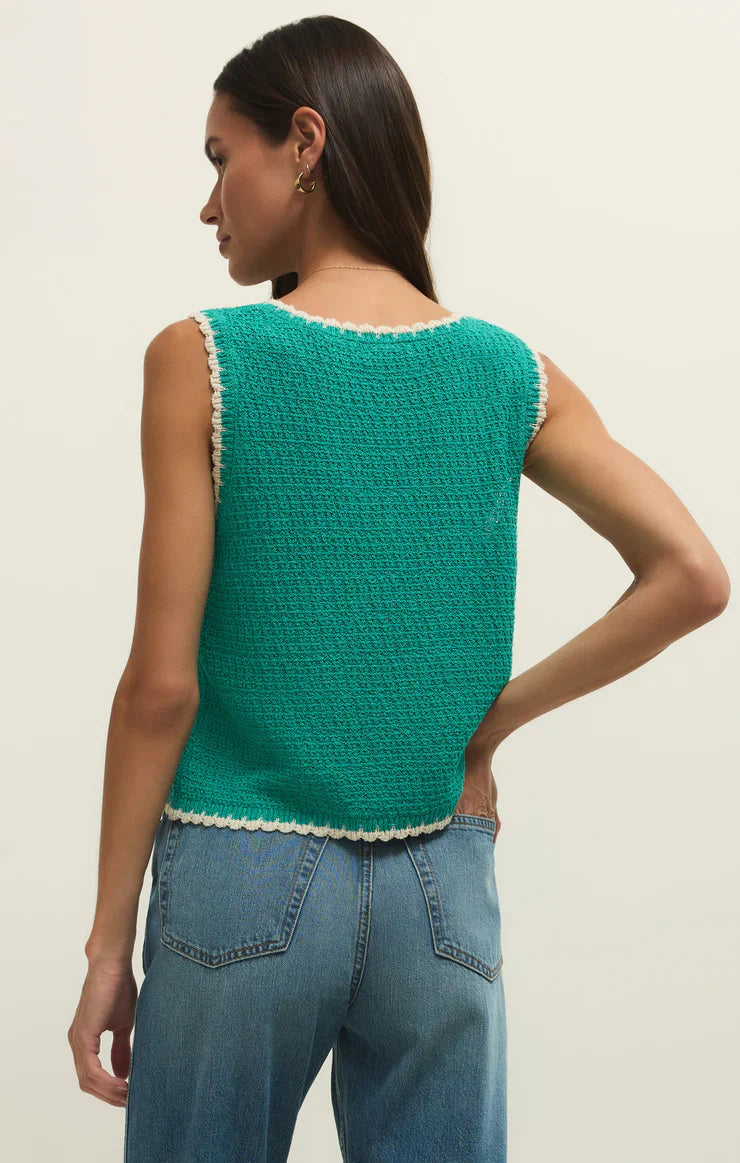 A woman with long dark hair stands with her back to the camera, wearing the Zertz YESSENIA WHIPSTICH TANK LA PALMA in green with contrast trim and blue jeans, her left hand resting on her hip.
