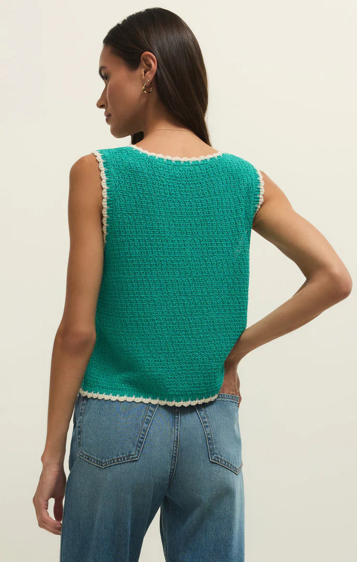 A woman with long dark hair stands with her back to the camera, wearing the Zertz YESSENIA WHIPSTICH TANK LA PALMA in green with contrast trim and blue jeans, her left hand resting on her hip.