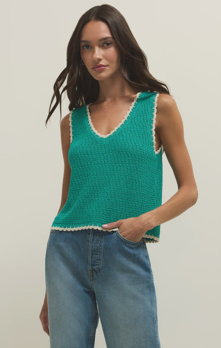 A woman wears the Zertz YESSENIA WHIPSTICH TANK LA PALMA, a sleeveless green yarn-blend top with contrast trim, paired with blue high-waisted jeans, standing against a plain light background with one hand in her pocket.