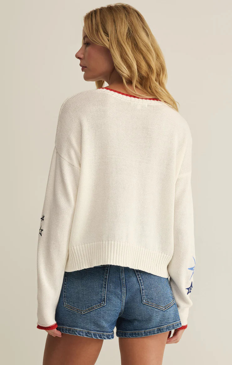 A woman with wavy blonde hair wears the Zertz SEAS THE DAY SIENNA SWEATER, a white cotton layer with red trim, paired with blue denim shorts. She stands with her back to the camera, looking left to showcase this perfect summer sweater.