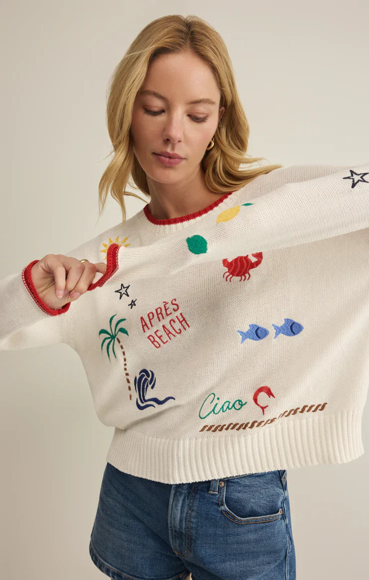 A woman wears the Zertz SEAS THE DAY SIENNA SWEATER—a white cotton sweater with colorful beach-themed designs and sayings—paired with blue denim shorts, making it the perfect summer layer.