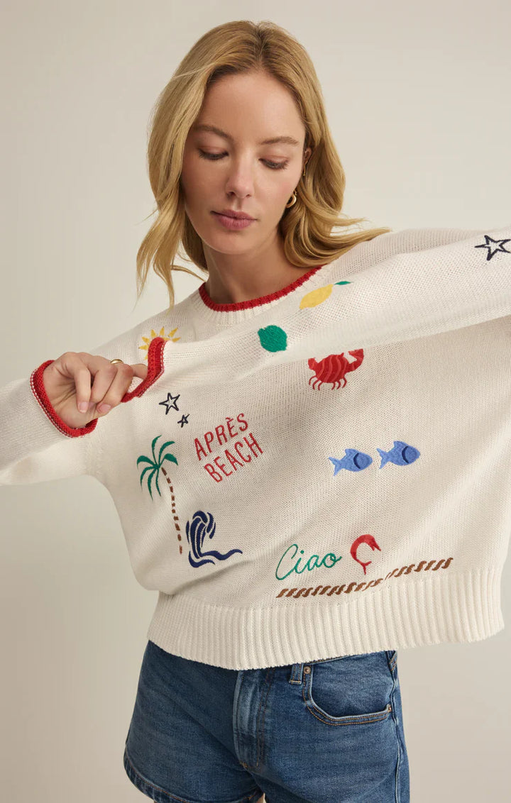 A woman wears the Zertz SEAS THE DAY SIENNA SWEATER—a white cotton sweater with colorful beach-themed designs and sayings—paired with blue denim shorts, making it the perfect summer layer.