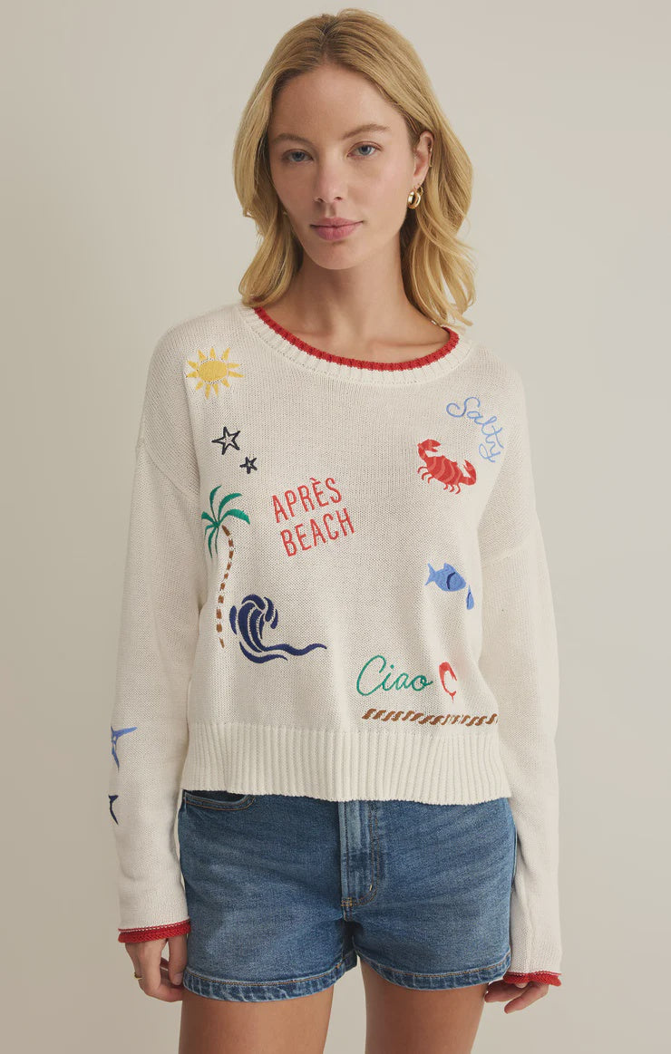 A woman with blonde hair models the Zertz SEAS THE DAY SIENNA SWEATER—white cotton with colorful beach-themed graphics and words—paired with denim shorts against a plain light background, making it the perfect summer layer.