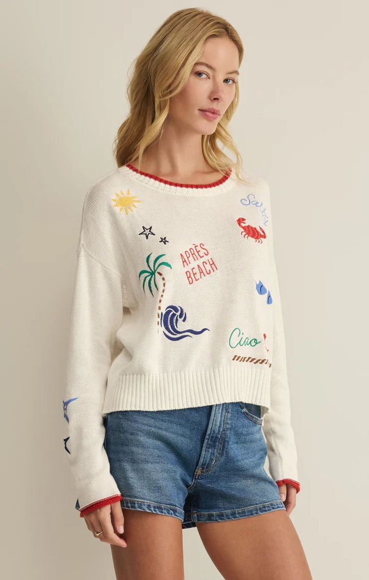 A woman with long blonde hair wears the Zertz SEAS THE DAY SIENNA SWEATER, a cotton beach layer with embroidered sun, crab, palm tree, and wave motifs plus beachy text, paired with denim shorts—perfect for summer.