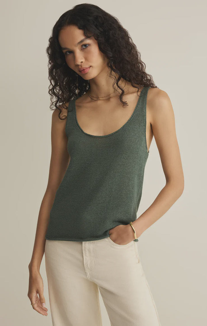 A person with long curly hair wears the Zertz PERCH SWEATER TANK in forest green and cream-colored pants, standing against a plain light background with one hand in their pocket.