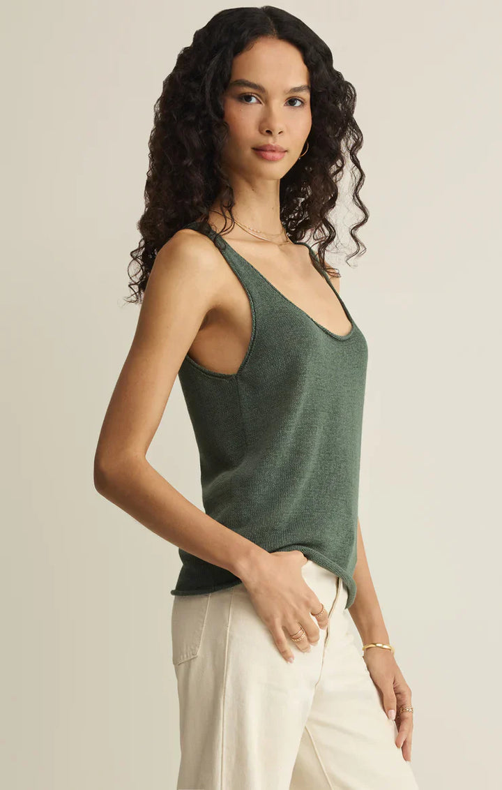 A woman with long curly hair wears the Zertz PERCH SWEATER TANK, a lightweight sleeveless top in dark green, paired with light beige pants. She stands sideways facing the camera, expression neutral, against a plain background.