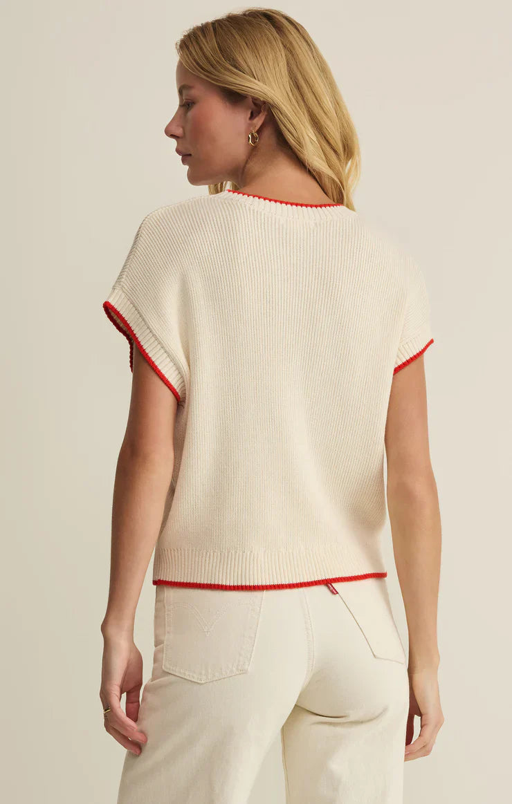 A woman with blonde hair faces away, wearing the Zertz SLEEVELESS PATRIOT SWEATER—a cream knit top with red trim—and high-waisted cream pants, set against a neutral background.