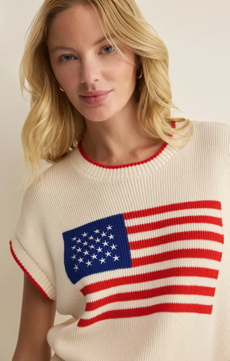 A woman with blonde hair wears the Zertz SLEEVELESS PATRIOT SWEATER, a cotton knit top featuring a large American flag on the front and red trim. She looks at the camera with a slight smile.