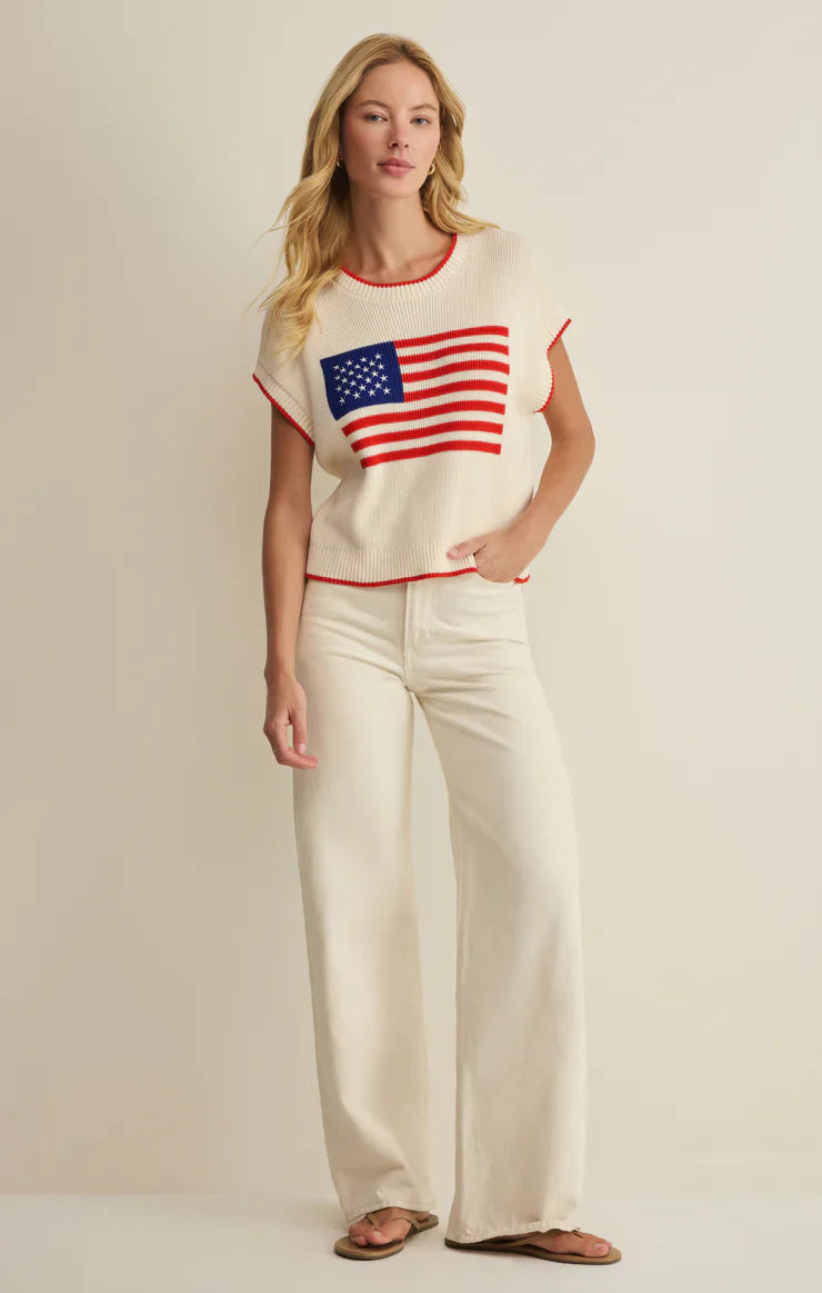 A woman with long blonde hair and a relaxed expression stands against a plain background wearing the Zertz SLEEVLESS PATRIOT SWEATER featuring an American flag, paired with cream wide-leg pants and sandals.