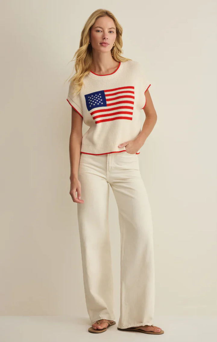 A woman with long blonde hair and a relaxed expression stands against a plain background wearing the Zertz SLEEVLESS PATRIOT SWEATER featuring an American flag, paired with cream wide-leg pants and sandals.
