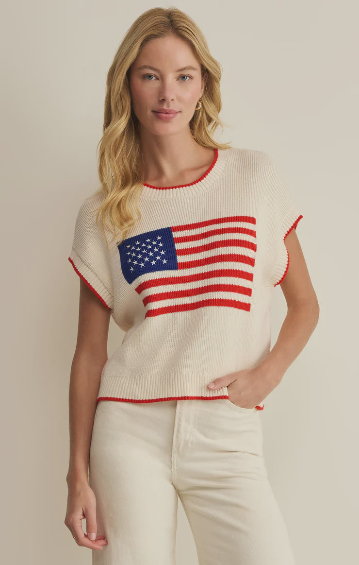 A woman with long blonde hair wears the Zertz SLEEVELESS PATRIOT SWEATER, a cream cotton knit with an American flag design, paired with cream pants. She stands against a neutral background, smiling softly with one hand in her pocket.