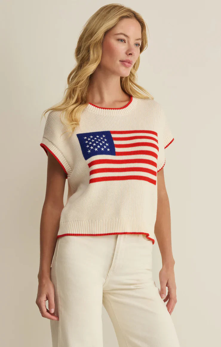A woman with long blonde hair wears the Zertz SLEEVELESS PATRIOT SWEATER, a lightweight cotton knit featuring a stylized American flag, paired with high-waisted white pants against a plain light background—perfect for summer.
