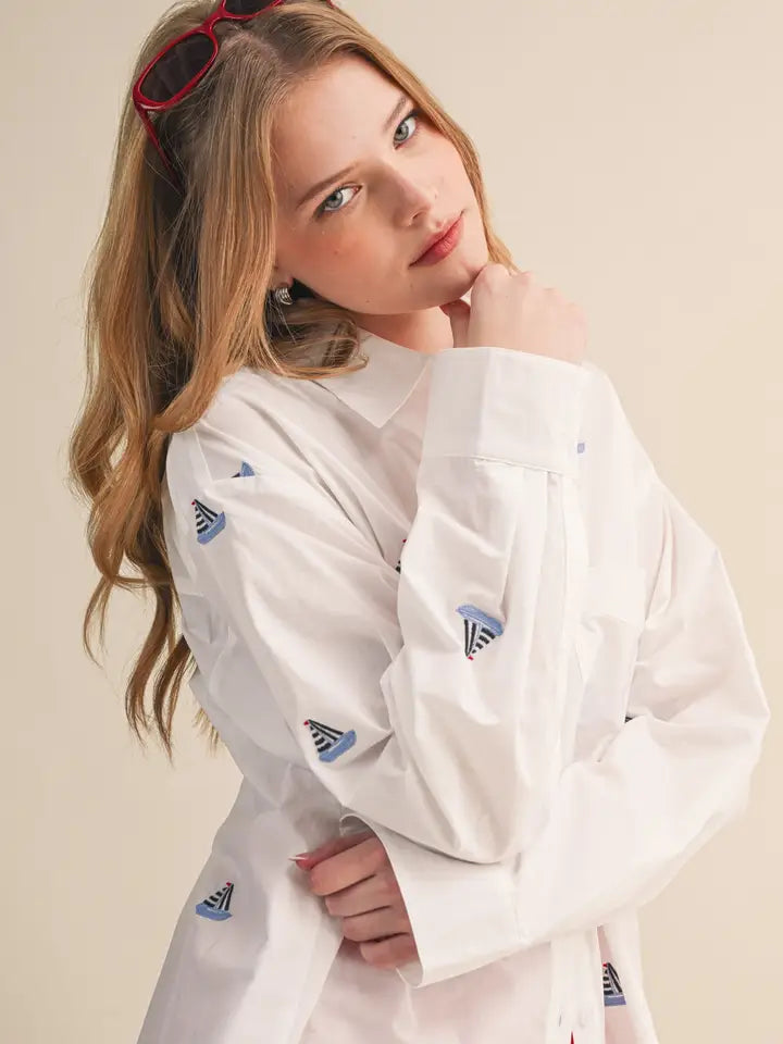 A young woman with long wavy blonde hair and red glasses wears the Zertz BOAT EMB TOP, a white shirt with small sailboat prints—a timeless staple—as she softly smiles at the camera, her arm raised near her chin against a plain backdrop.
