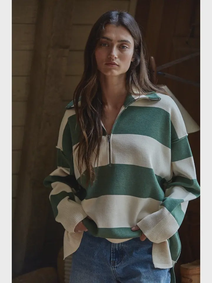 A woman with long brown hair models the Zertz FONTAINE HALF ZIP SWEATER, a green and white striped knit with loose sleeves, paired with blue jeans. She stands indoors against a wooden wall.