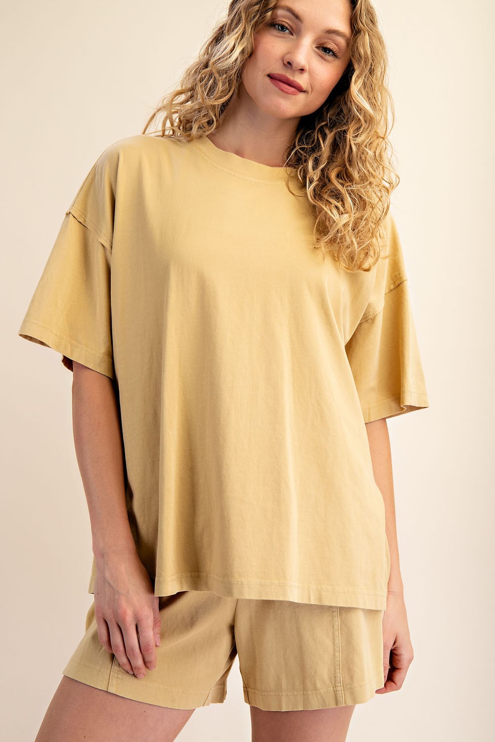 A woman with curly blonde hair is wearing The Mellow Yellow Set by Loca on Main—an everyday lounge set featuring a loose-fitting, mineral-washed tee and relaxed fit shorts. She stands smiling softly against a neutral background.