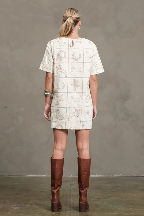 A woman with blonde hair in a low ponytail faces away, wearing the Zertz WESTERN EMB MINI DRESS in cream with printed designs and tall brown boots, standing on a concrete floor against a plain gray wall.