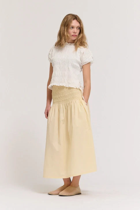 A woman with light brown hair and a neutral expression models the Zertz SMOCKED WAIST SKIRT, pairing it with a white short-sleeve ruched top and beige slip-on shoes against a plain background. The skirt is high-waisted, light beige, and features pockets.