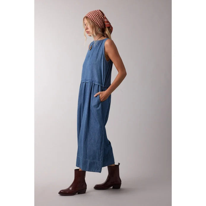 A woman stands in profile against a plain light background, wearing the Zertz SLVLESS MAXI DRESS, brown ankle boots, and a red patterned headscarf, with one hand in her pocket.