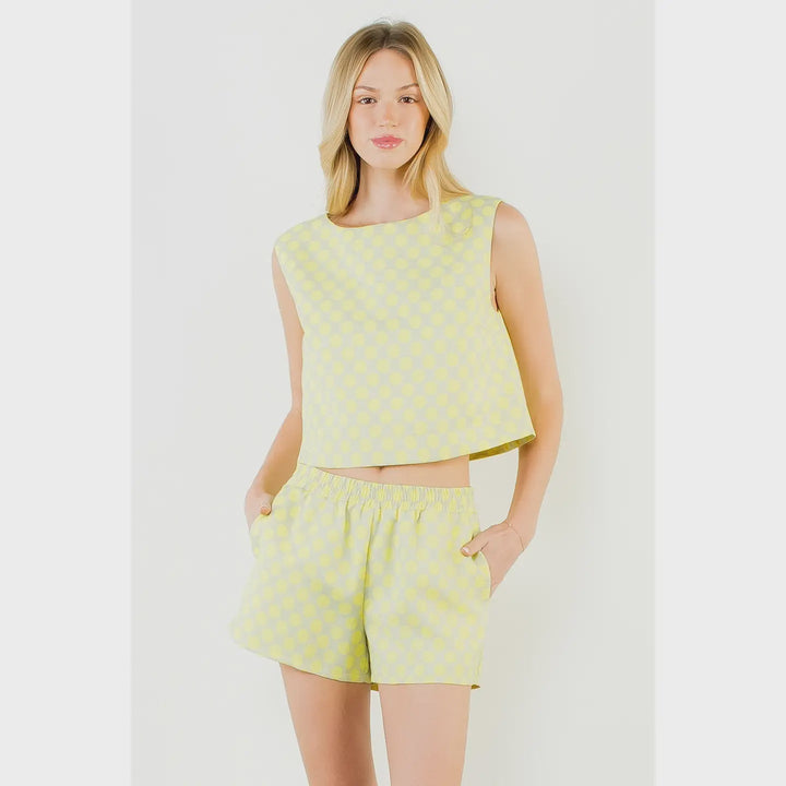A woman stands against a white background wearing the Zertz POLKA DOT TOP, a sleeveless yellow and light blue checkered crop top with matching shorts. Her hands are in her pockets as she looks at the camera with a relaxed expression.