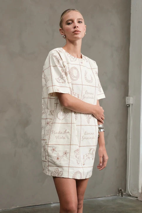 A woman wears the Zertz WESTERN EMB MINI DRESS—a short-sleeved, cream artisan mini dress featuring brown line art and text—standing against a gray wall and looking confidently at the camera with one arm on her opposite elbow.