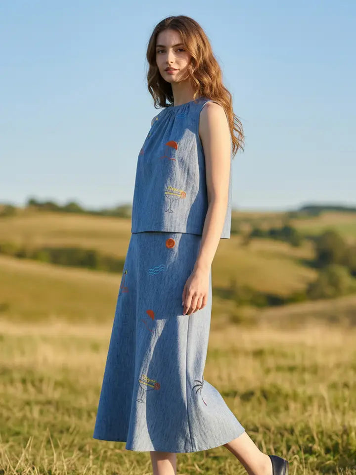 A woman in a sleeveless blue SUNSET EMBROIDERY SET by Zertz with subtle embroidery stands in a sunlit grassy field, rolling hills behind her.