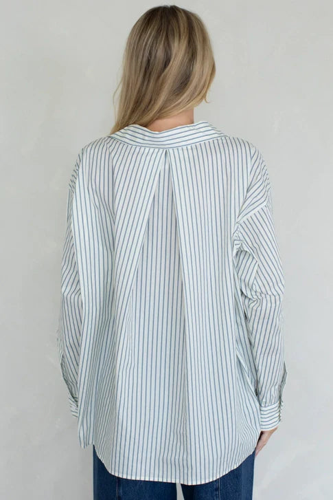 A person with long blonde hair is shown from the back, wearing the Zertz STRIPED OVER SIZED BUTTON UP TOP— a loose-fitting, long-sleeve white shirt with vertical blue stripes— and blue jeans against a plain light background.