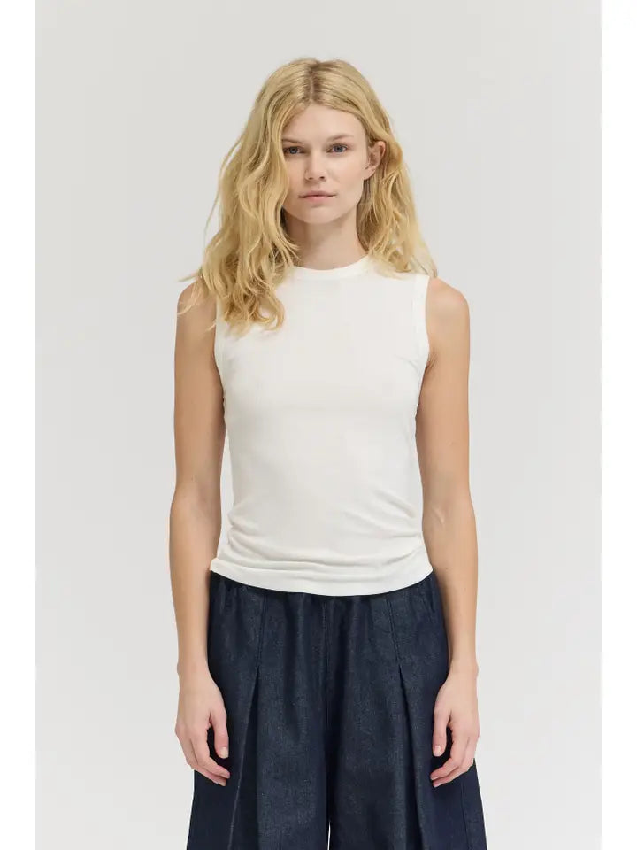 A woman with long blonde hair models the Zertz SHEER TANK TOP, a sleeveless lightweight white top, paired with dark wide-leg pants. She stands against a plain light background, facing the camera.