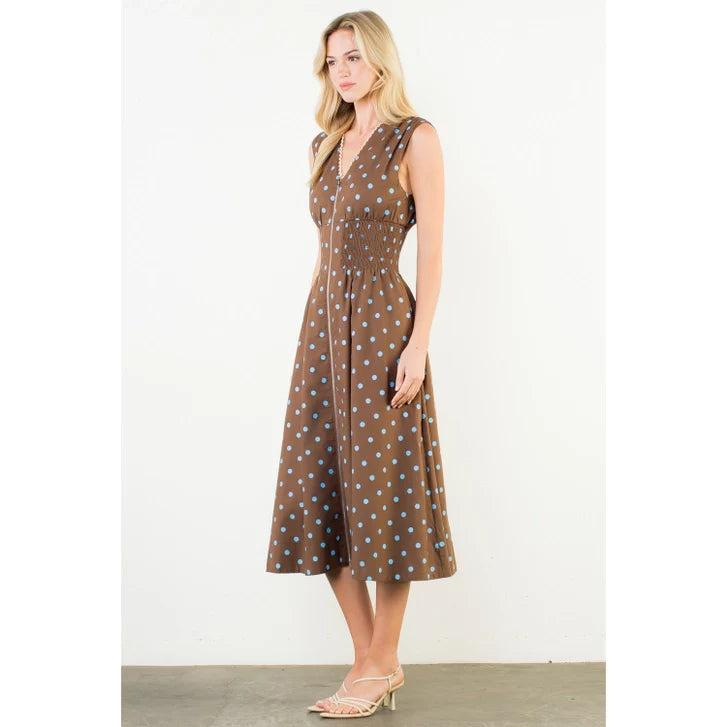 A woman with long blonde hair wears the Zertz SLVLESS POLKA DOT MIDI DRESS, a brown sleeveless dress with blue dots, fitted waist, and front buttons. She pairs it with beige heeled sandals against a plain white background.
