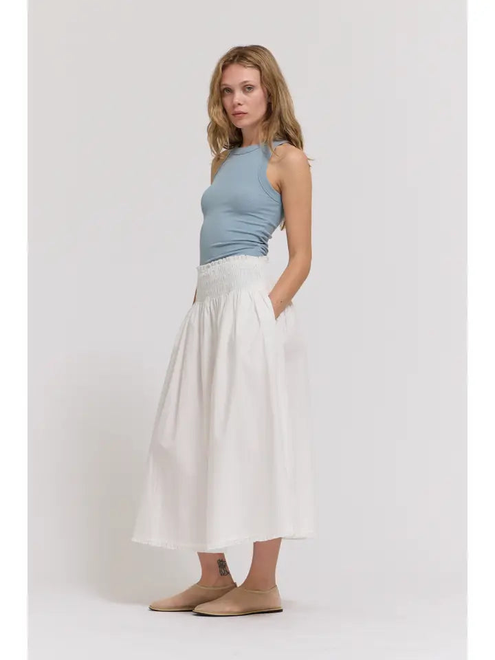 A woman stands against a white background, wearing a light blue sleeveless top and the Zertz SMOCKED WAIST SKIRT—a high-waisted white midi skirt with eyelet trim and pockets—paired with beige flats, looking to the side with a relaxed expression.