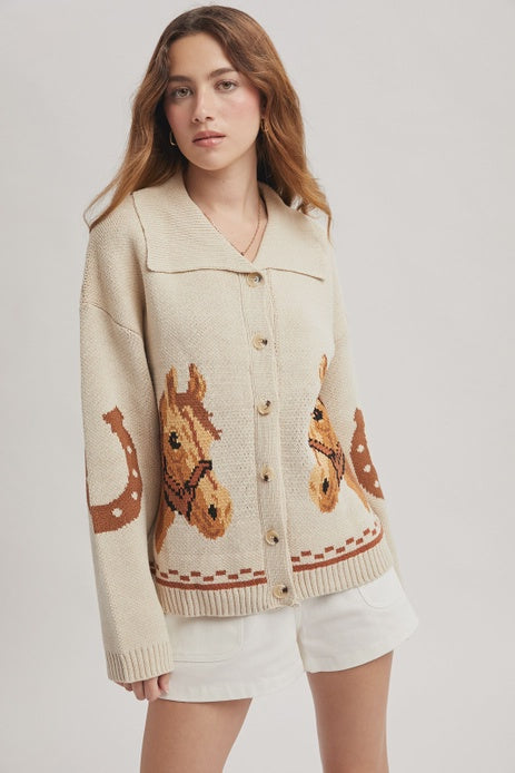 A woman with wavy brown hair models the Zertz HORSE GRAPHIC BUTTON CARDIGEN, a western-inspired long-sleeve piece with horse and horseshoe motifs, styled with white shorts against a plain light background.