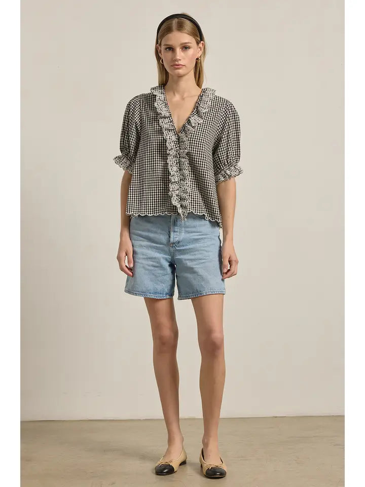 A woman poses against a plain backdrop in Zertz’s GINGHAM LACE RUFFLE BLOUSE, paired with light blue denim shorts, a black headband, and beige flats with black toe caps.