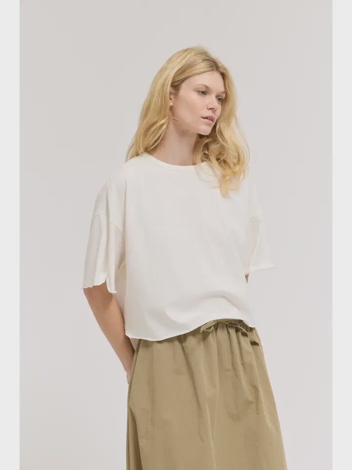 A woman with long blonde hair wears the Zertz LOOSE LONG SLEEVE TEE and an olive green skirt, standing against a plain light background and looking slightly to the side with a neutral expression.