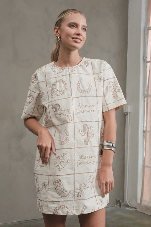 A woman stands by a window, smiling slightly, her hair in a low ponytail, wearing the Zertz WESTERN EMB MINI DRESS featuring cream fabric with brown Western-themed embroidery.