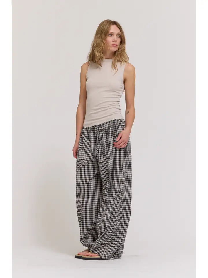 A woman with wavy, light brown hair and a relaxed expression wears the Zertz SHEER TANK TOP against a plain white background, paired with loose black and white checkered pants and sandals.